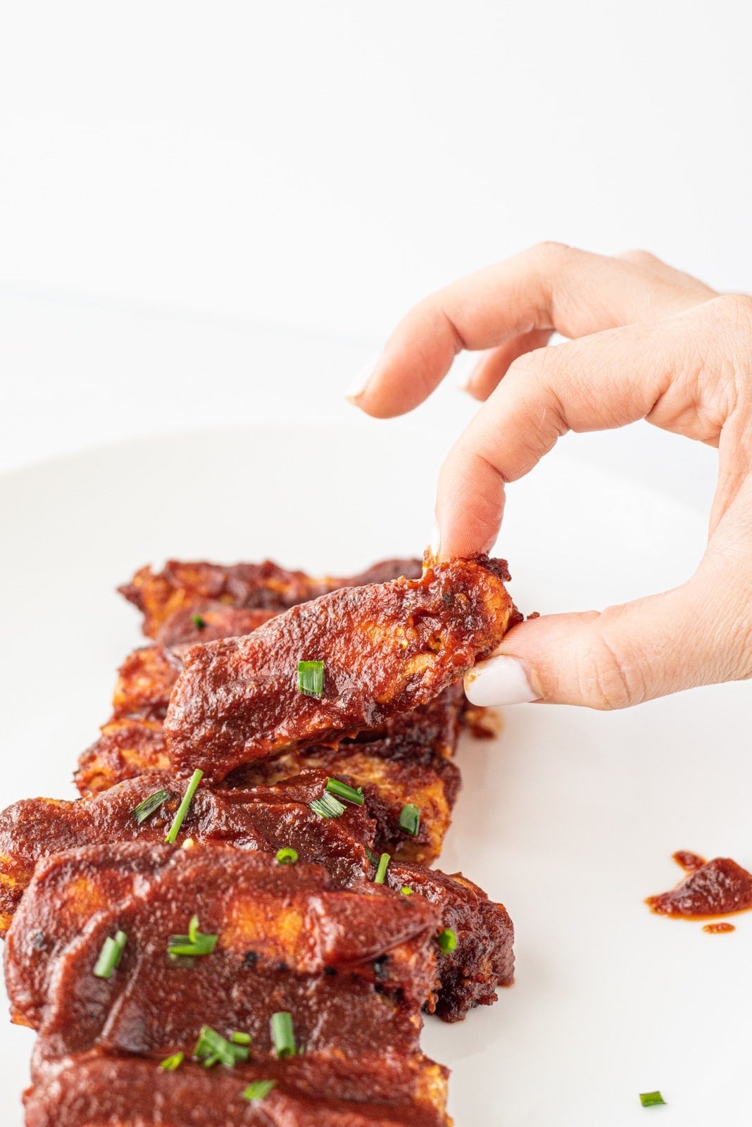 A white plate with vegan tempeh ribs on it and a white hand picking one up.