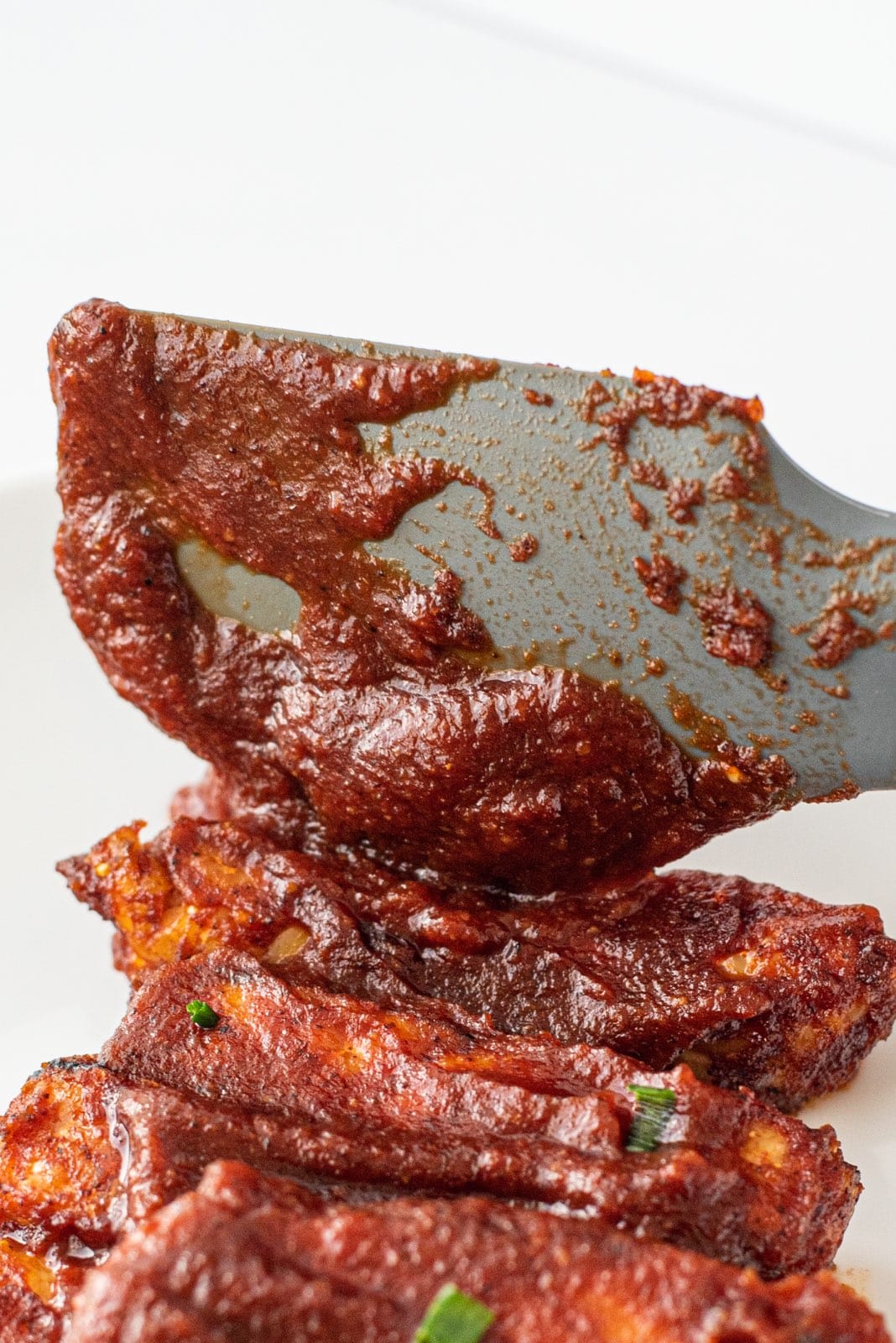 Vegan BBQ Tempeh Ribs on a white plate with a rubber spatula spreading BBQ sauce on them.
