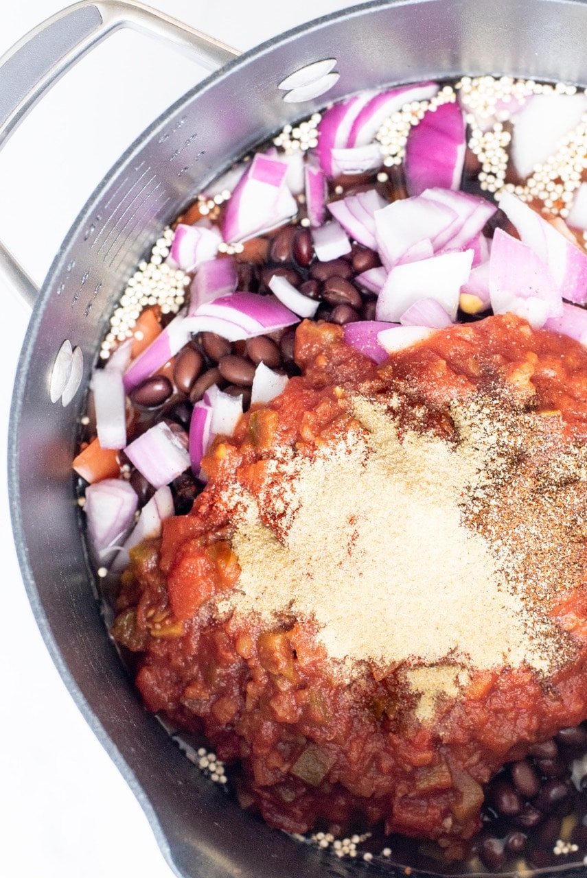 A close up of a silver pot filled with quinoa, water, spices, bell pepper, red onion, black beans, and corn.
