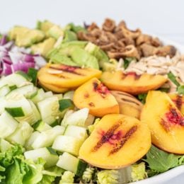 A white bowl filled with a salad topped with grilled peaches, chopped dried figs, diced cucumber, avocado cubes, shaved almonds, and diced red onion.