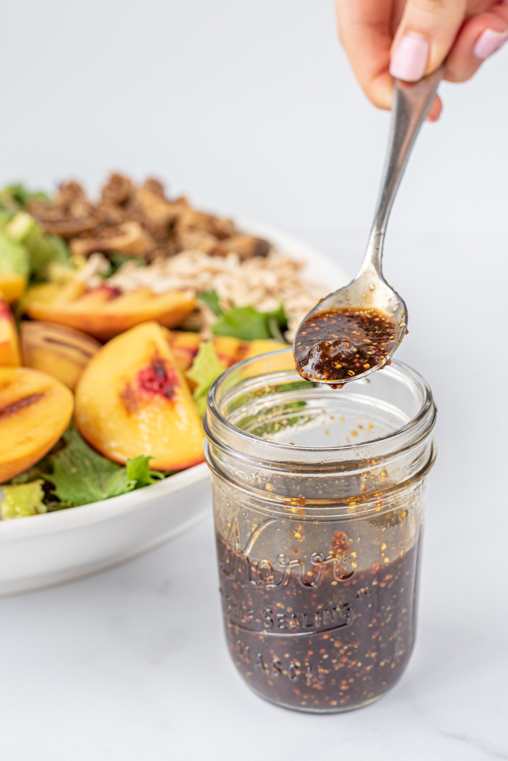 A small glass jar filled with balsamic dressing and a silver metal spoon over top the jar holding a spoonful of dressing. In the background is a salad.