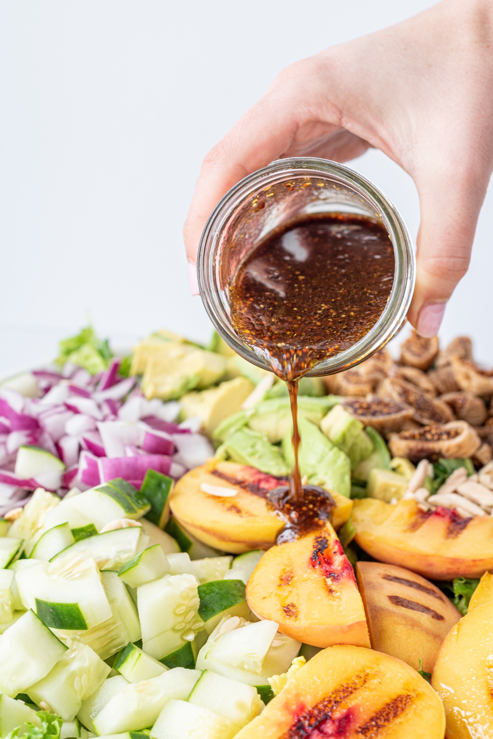 A white hand holding a jar filled with balsamic dressing pouring overtop a salad topped with peaches, red onion, cucumber, and avocado.