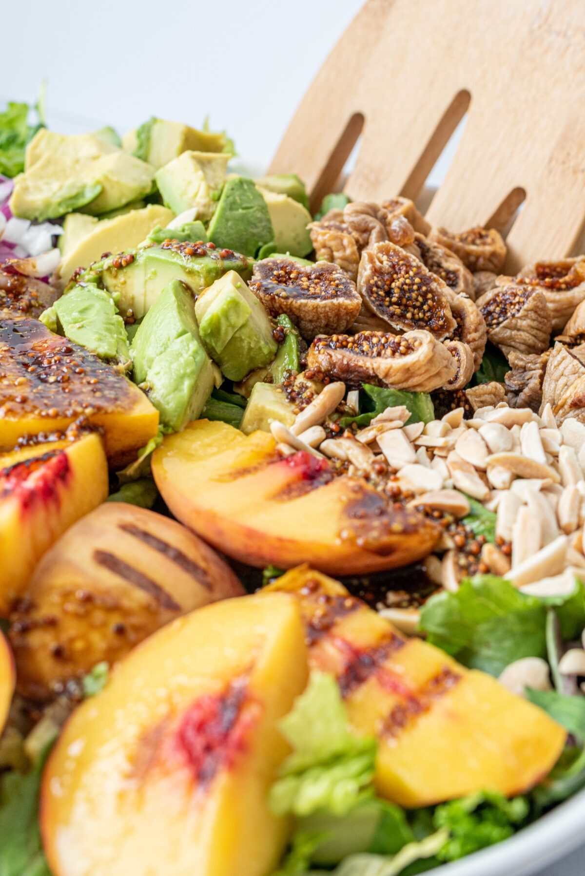 A salad topped with grilled peaches, dried figs, sliced almonds, avocado cubes, and a set of wooden tongs.
