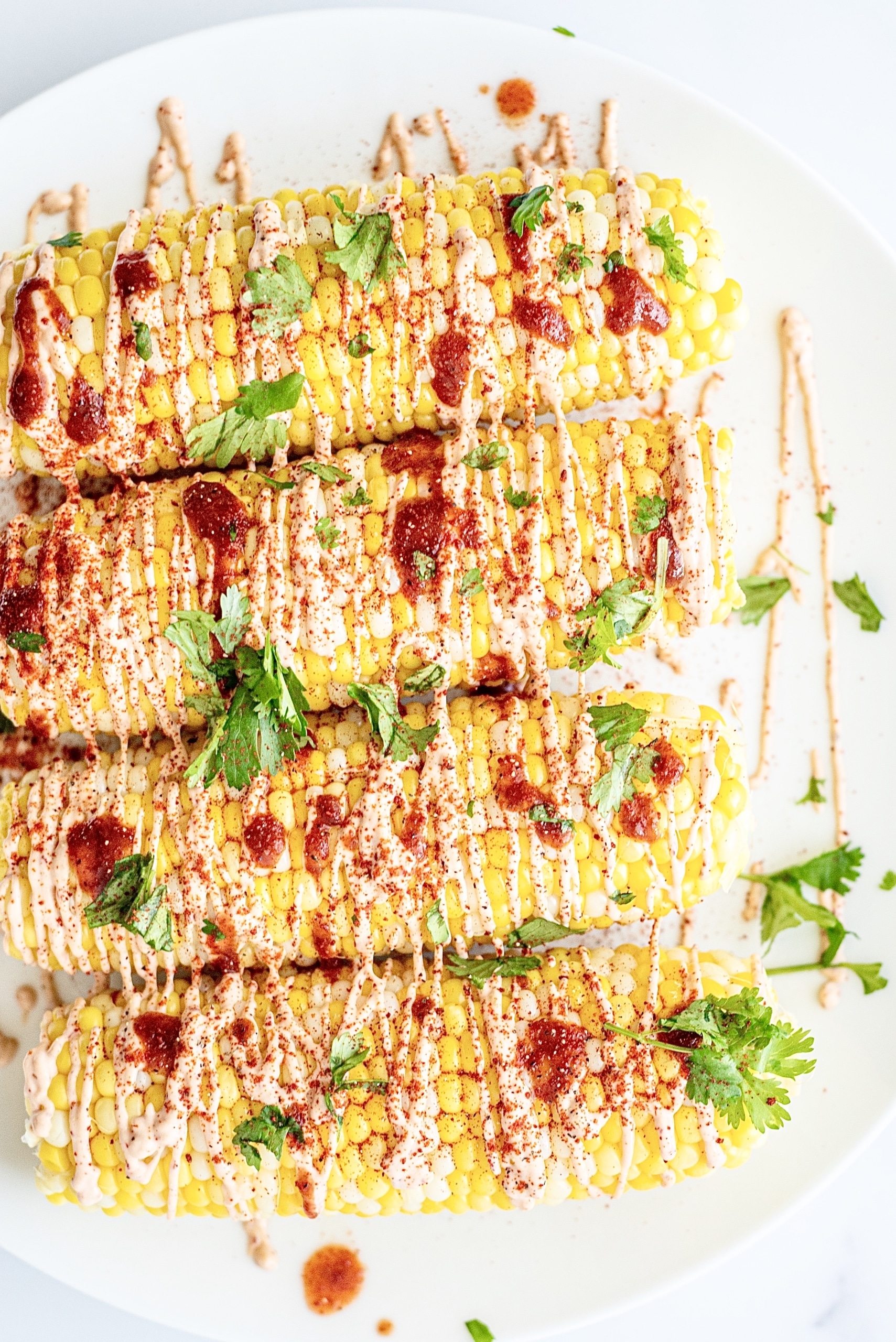 A white plate with four ears of corn topped with hot sauce, cilantro, and spicy aioli.