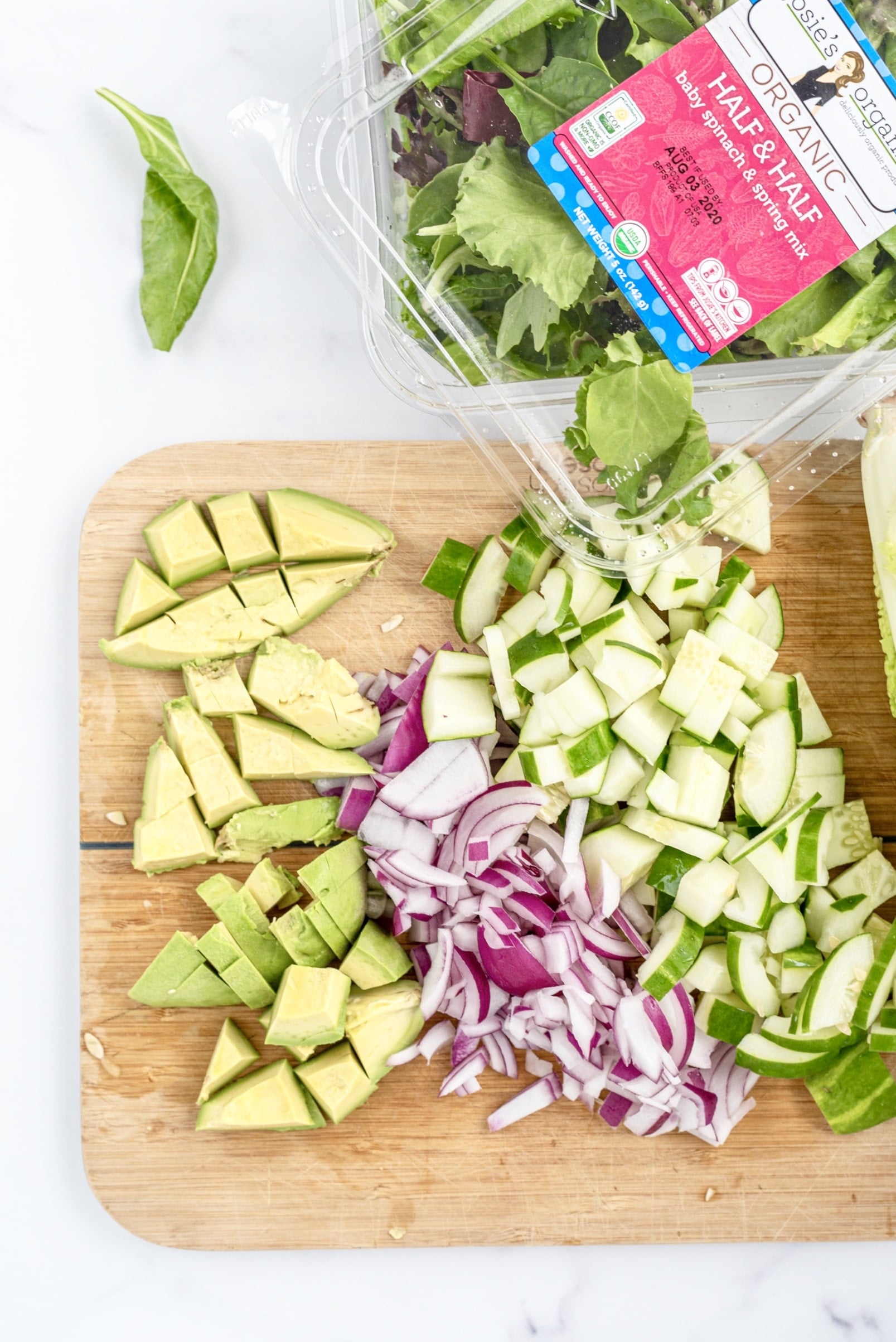 A wooden cutting board filled with chopped avocado, red onion, and cucumber, and a clear plastic container of spring mix.