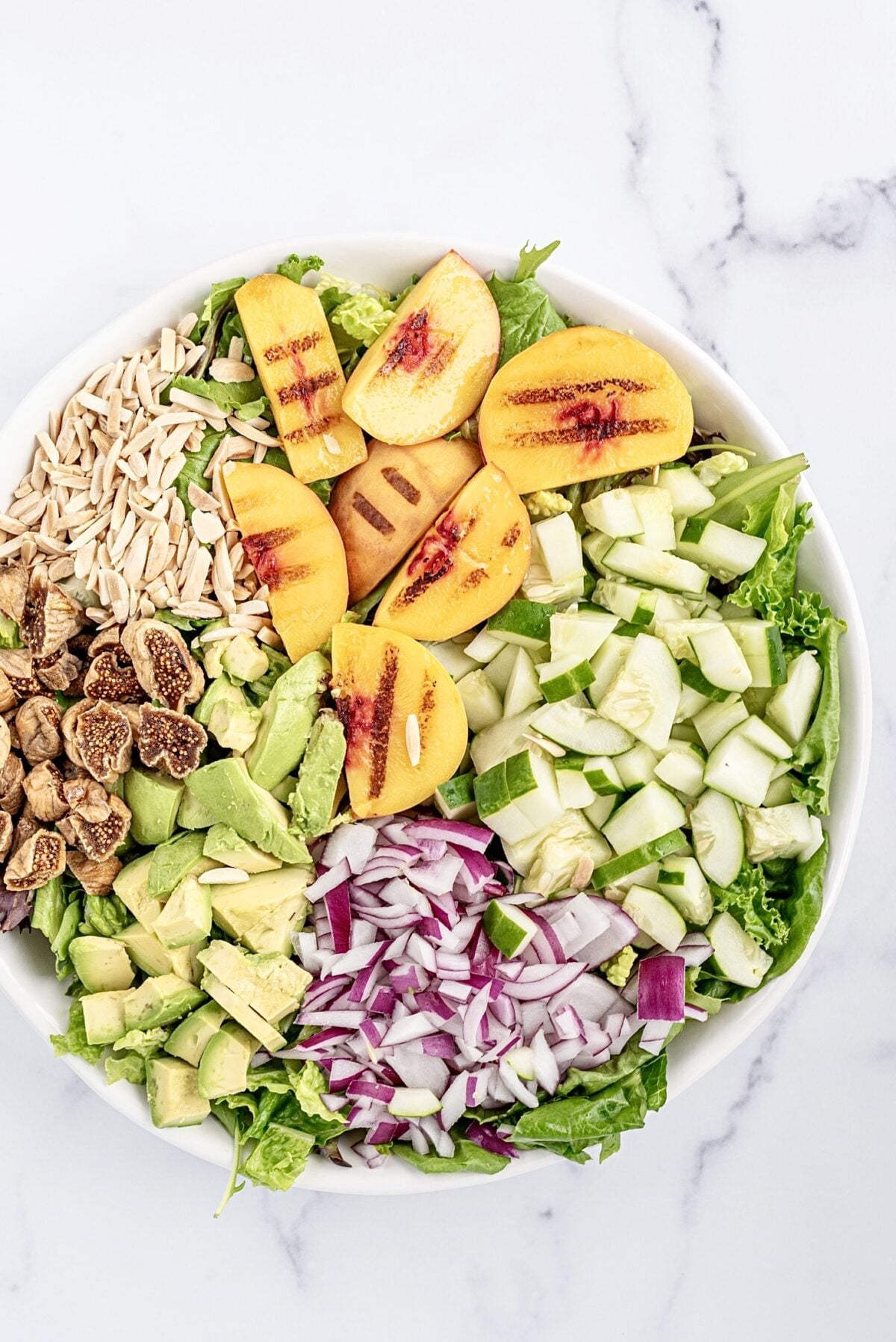 A white bowl filled with a salad topped with grilled peaches, chopped dried figs, diced cucumber, avocado cubes, shaved almonds, and diced red onion.