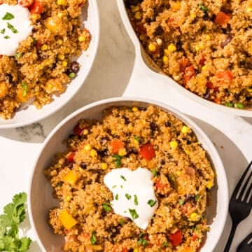 Two white bowls of one pot mexican quinoa garnished with sour cream and cilantro. Beside the bowls is a sprig of fresh cilantro, two black forks, and a large pot filled with remaining quinoa.