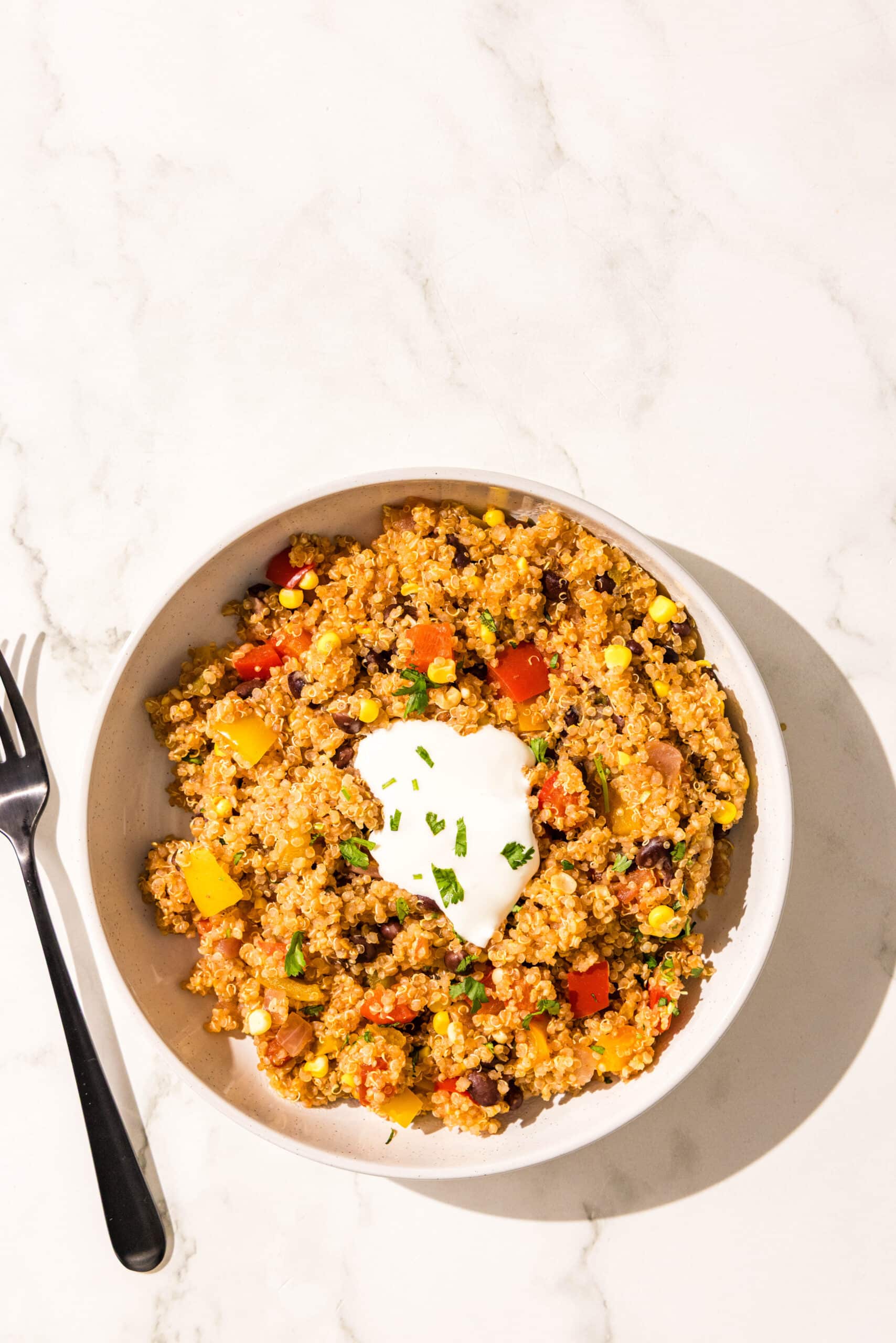 A white bowl filled with a serving of one pot mexican quinoa garnished with sour cream and cilantro. To the left of the bowl is a black metal fork.