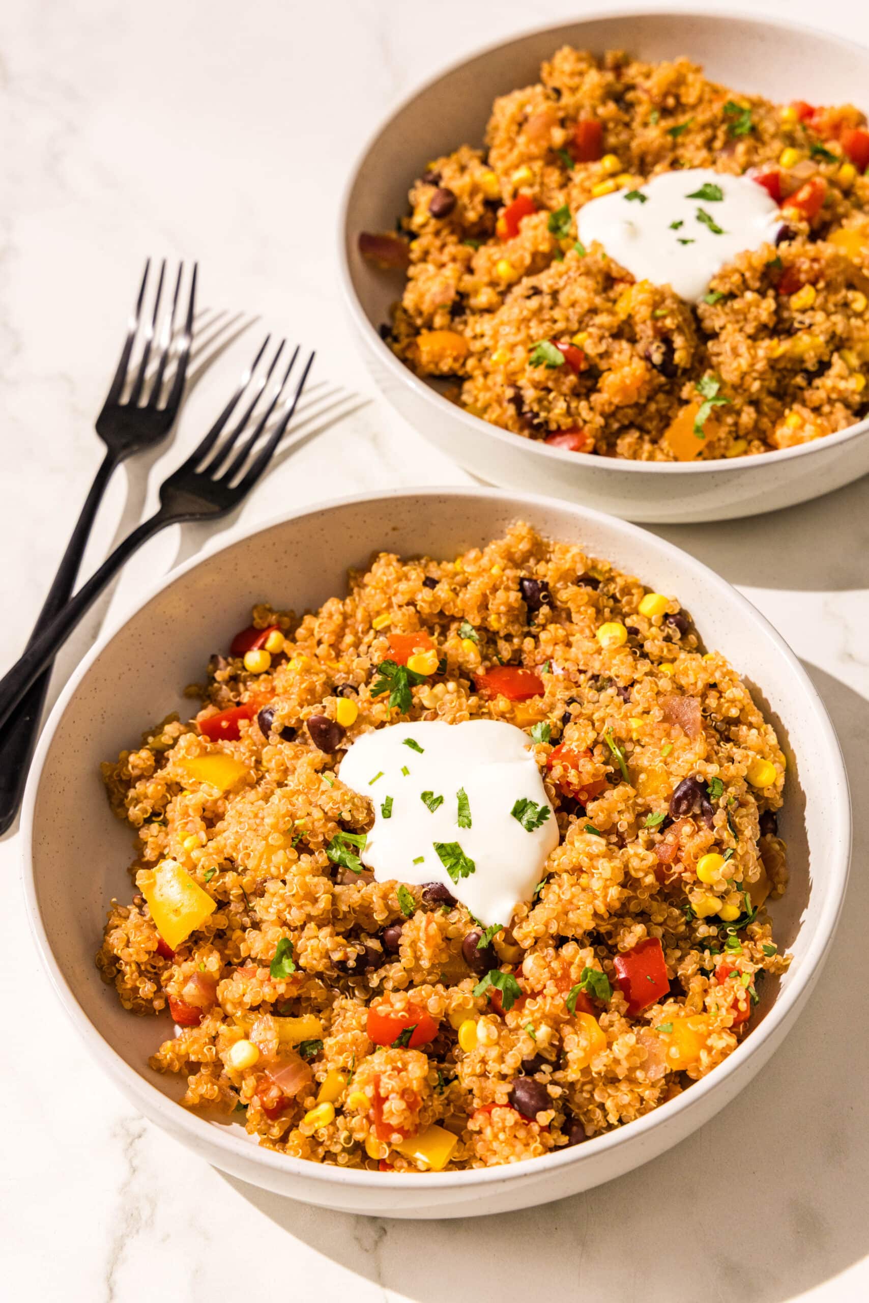 Two white bowls of one pot mexican quinoa topped with sour cream and cilantro. Beside the bowls is two black metal forks.