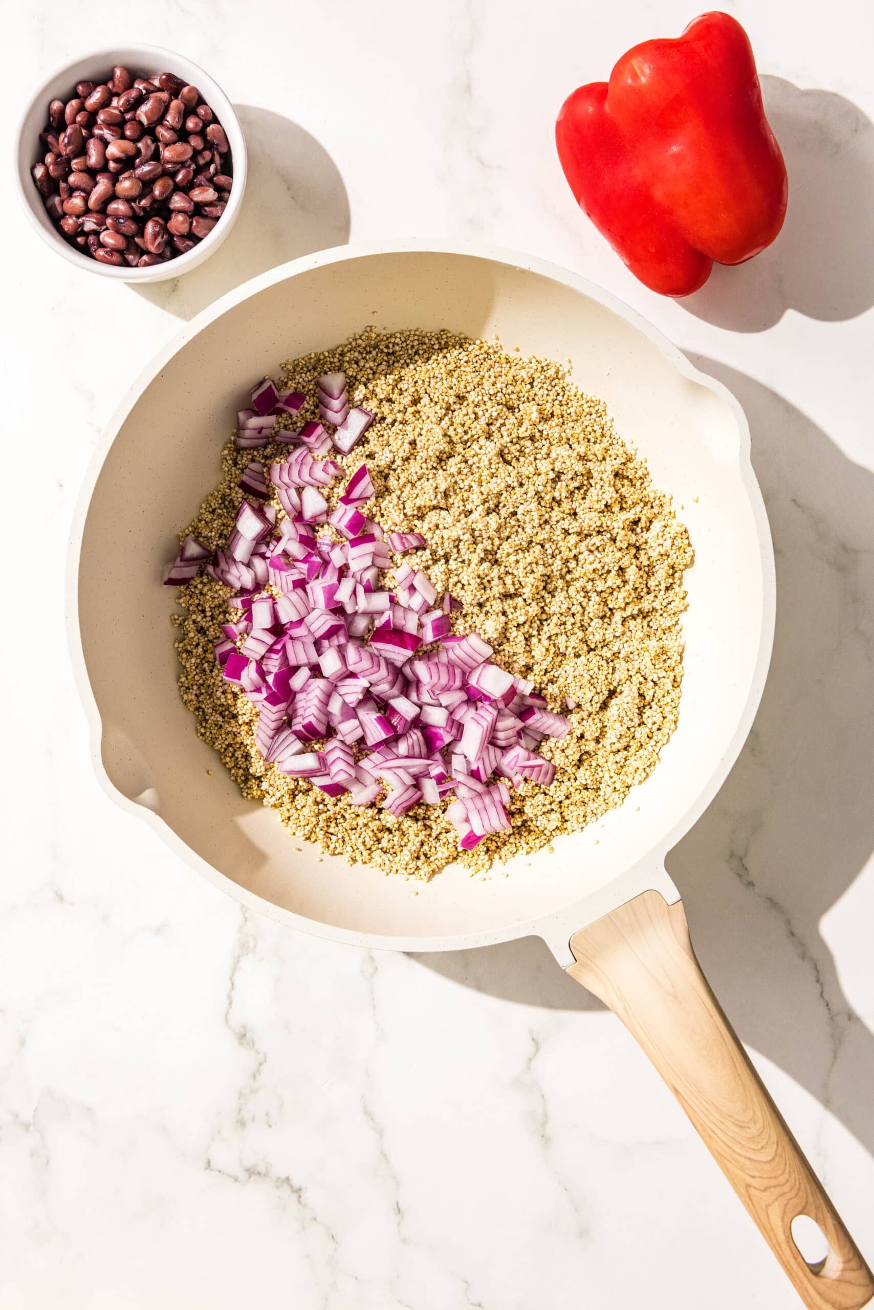 A white pot filled with quinoa and red onion. Beside the pot is a red bell pepper and a bowl of black beans.