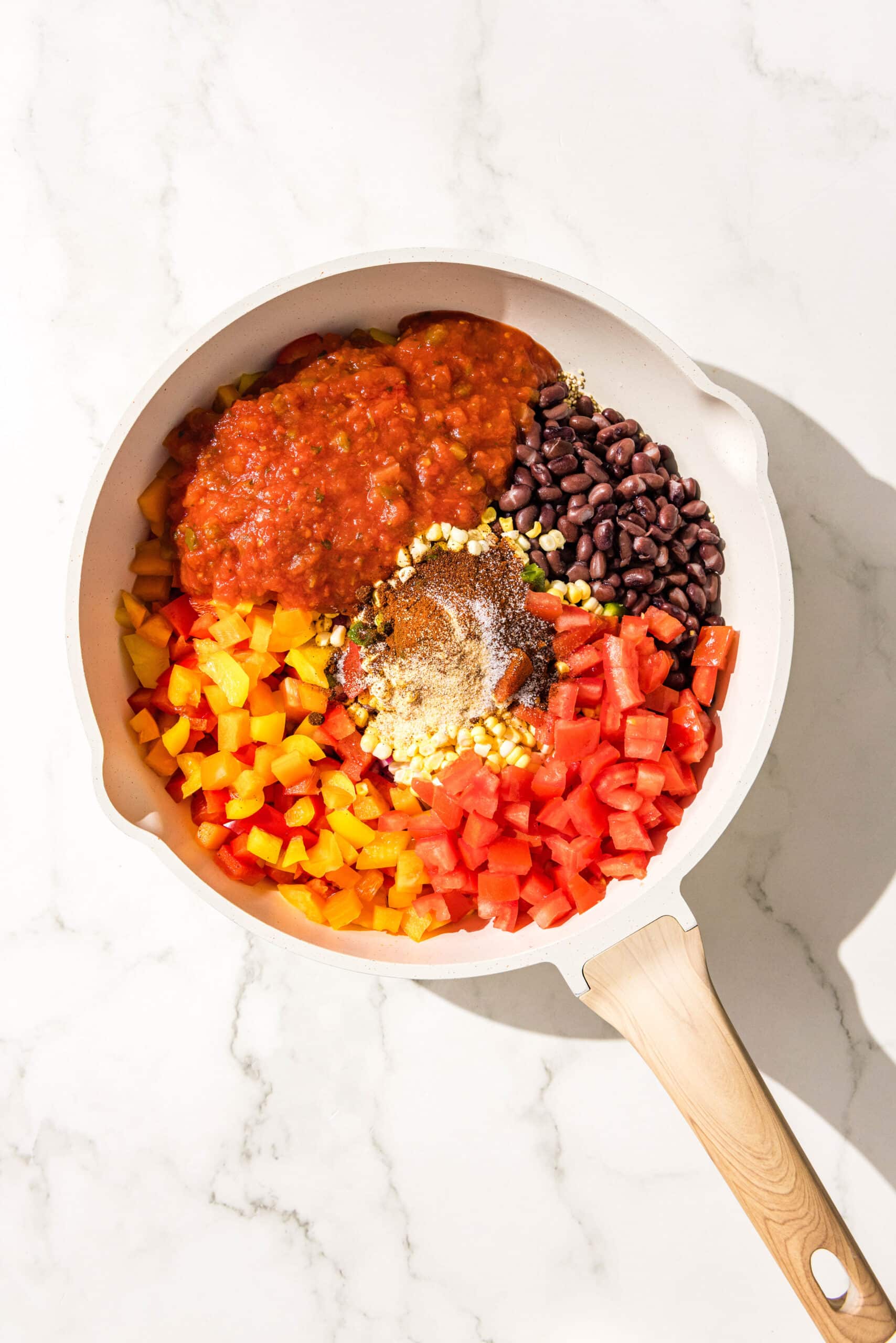 A large white pot of dry quinoa, red bell pepper, red onion, black beans, tomatoes, salsa, corn, and spices.