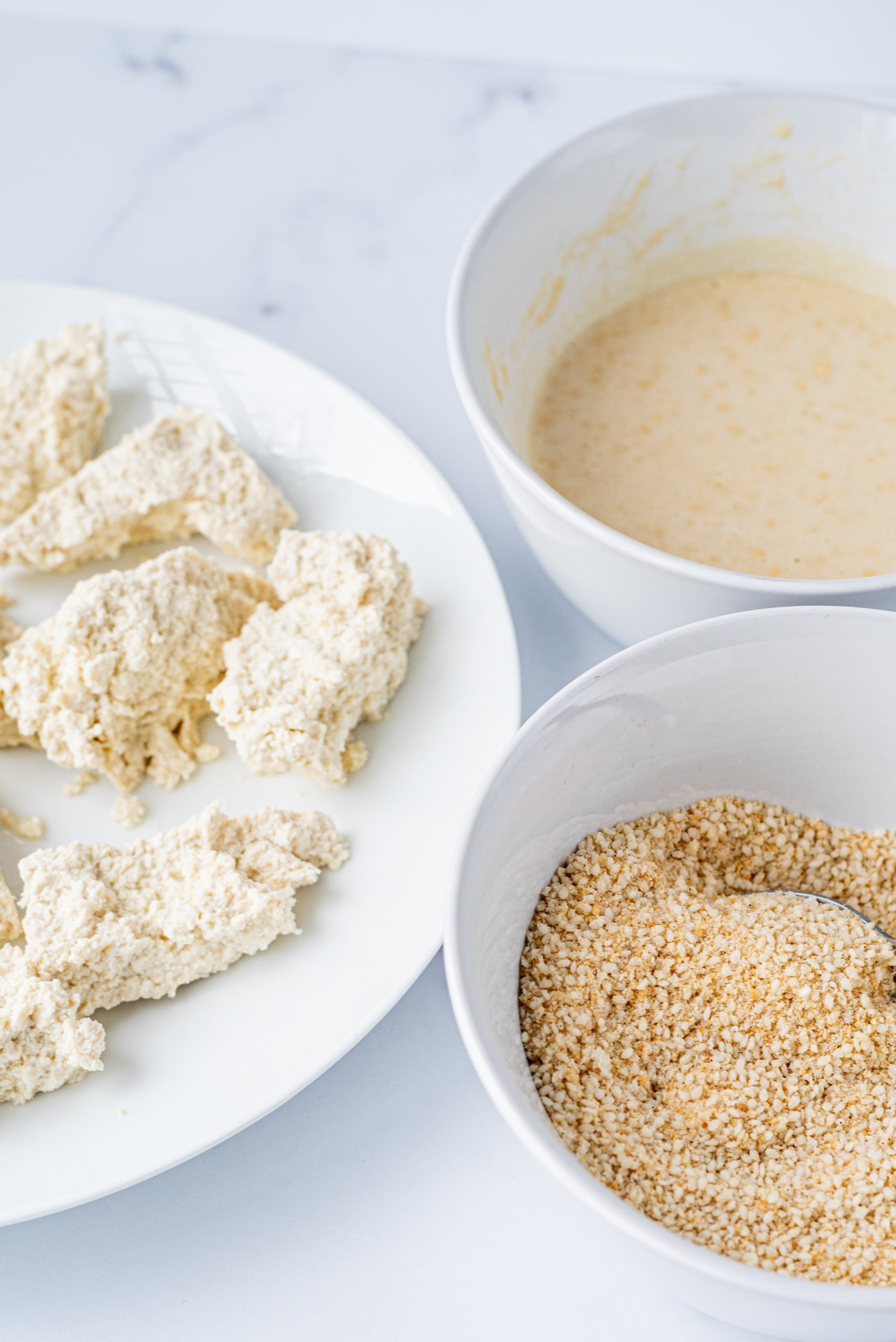 A white plate with tofu nuggets on it and two small bowls of breadcrumbs and flour.