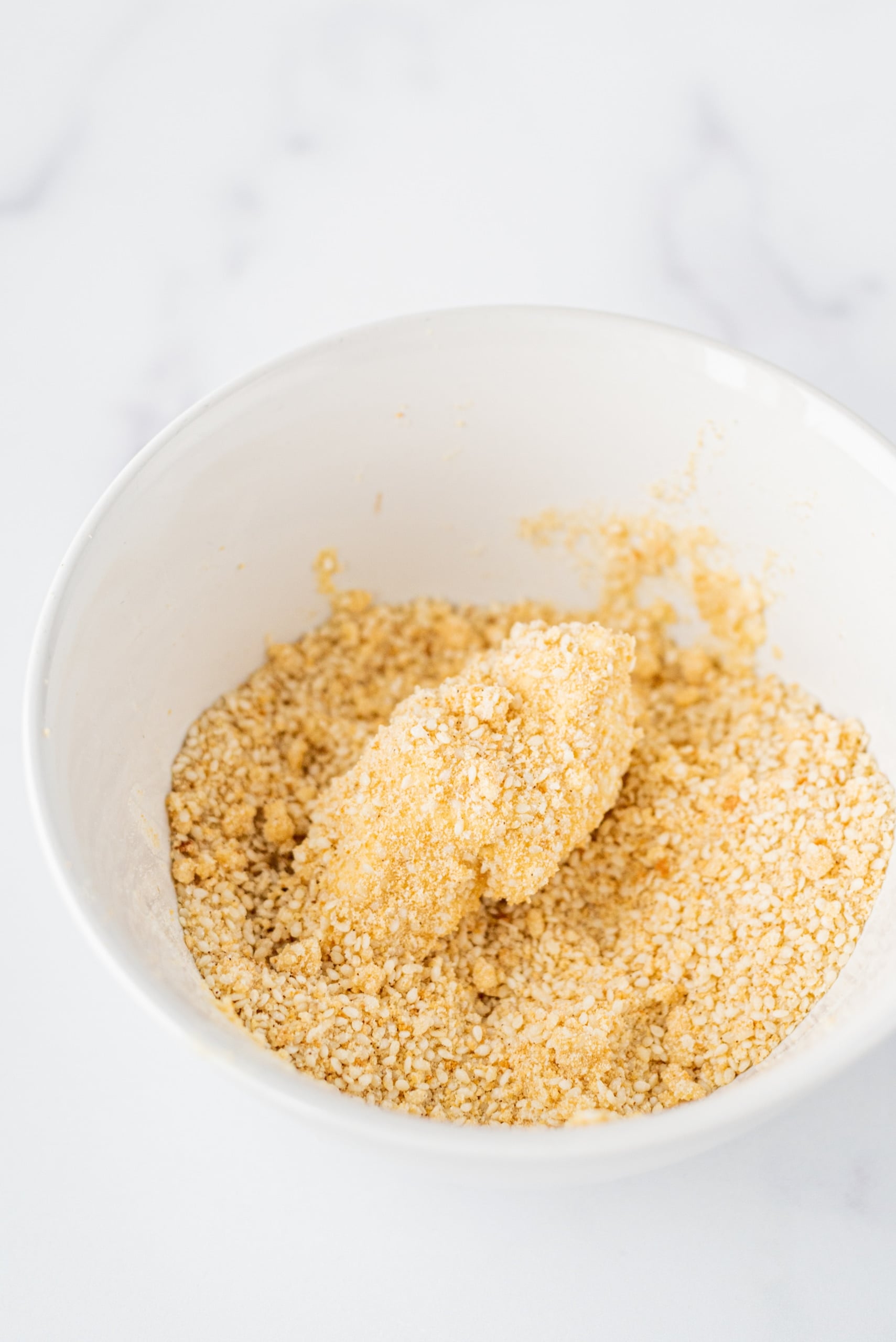 A white bowl of breadcrumbs with a tofu nugget rolled in the crumbs.