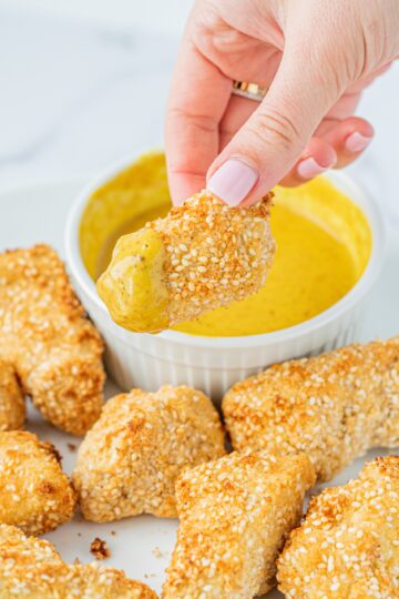 A white plate of vegan tofu tenders and a bowl of honey mustard and a white hand holding a nugget covered in sauce.