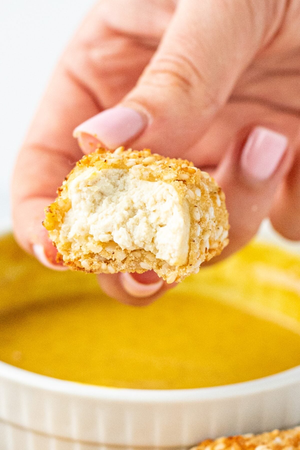 A white hand holding a bitten into tofu tender and a bowl of honey mustard dipping sauce.
