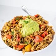 A large white glass bowl filled with vegan taco pasta salad topped with a scoop of avocado cilantro sauce in the center.