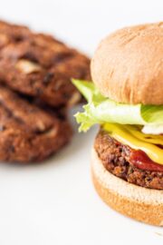 Two black bean burger patties on a white plate next to a burger with a black bean patty on it, mayonnaise, mustard, a slice of tomato, and a big piece of lettuce.