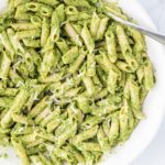 A white bowl filled with green pasta and a silver fork sticking out.