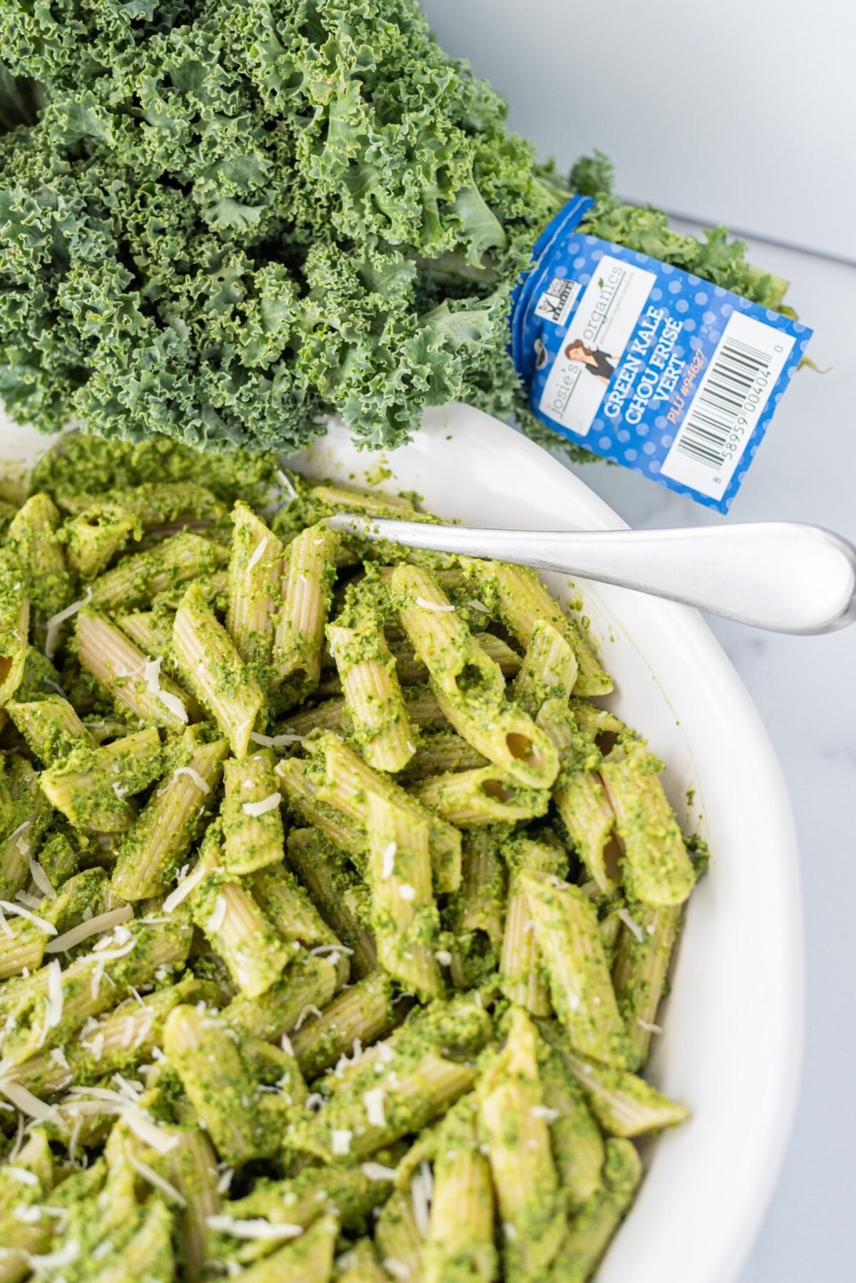 A white bowl filled with pesto pasta and a silver fork sticking out. Beside the bowl is a bundle of fresh kale.