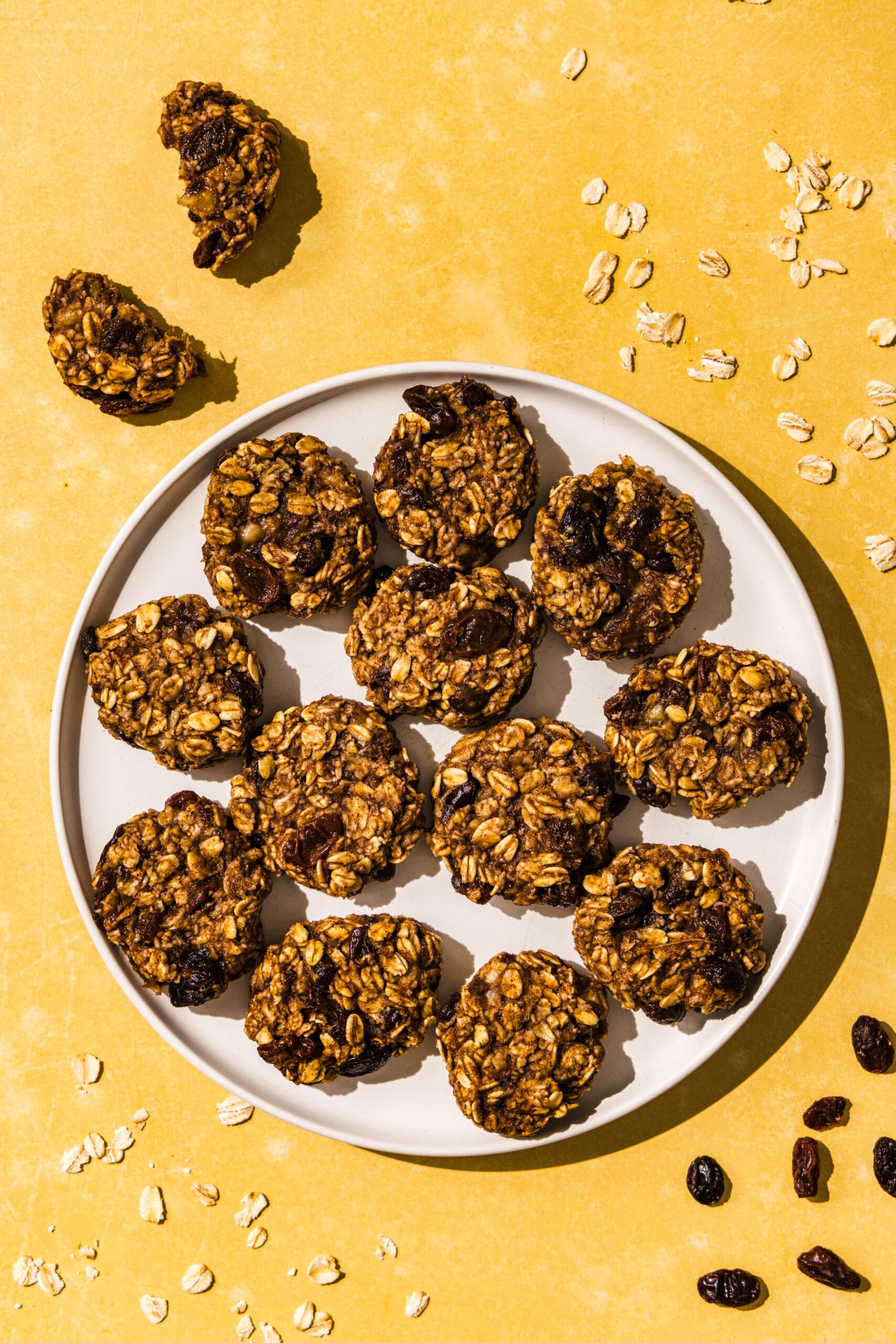 A white plate with 3-ingredient banana oatmeal bites on it. Around the plate is one oatmeal bite broken in half and then sprinkled raisins and oats on the counter.