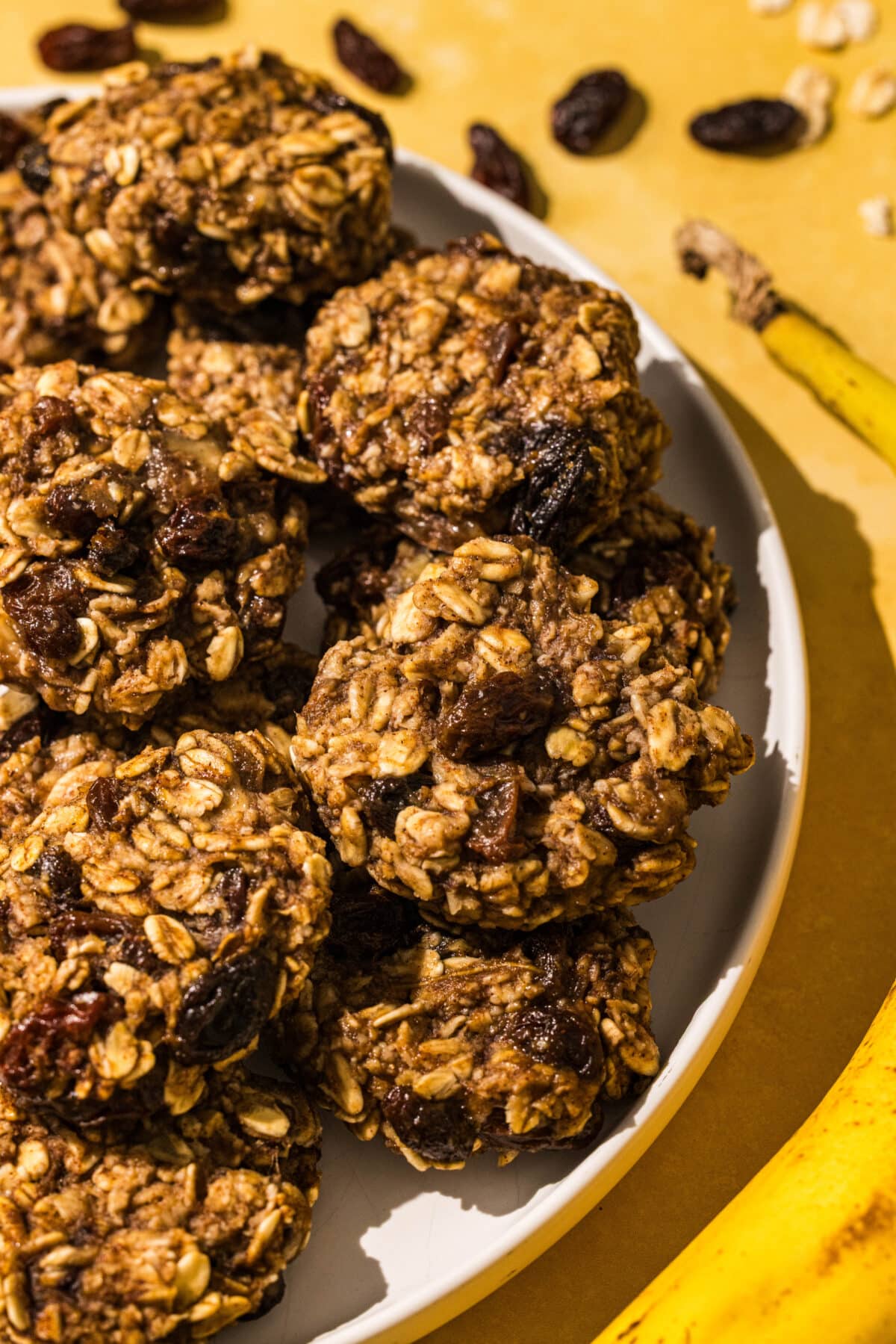 Close up of the three ingredient banana oatmeal bites stacked up on a white plate, with only half of the plate showing from the lefthand side. To the right of the plate is a banana and some raisins and oats sprinkled on the counter.