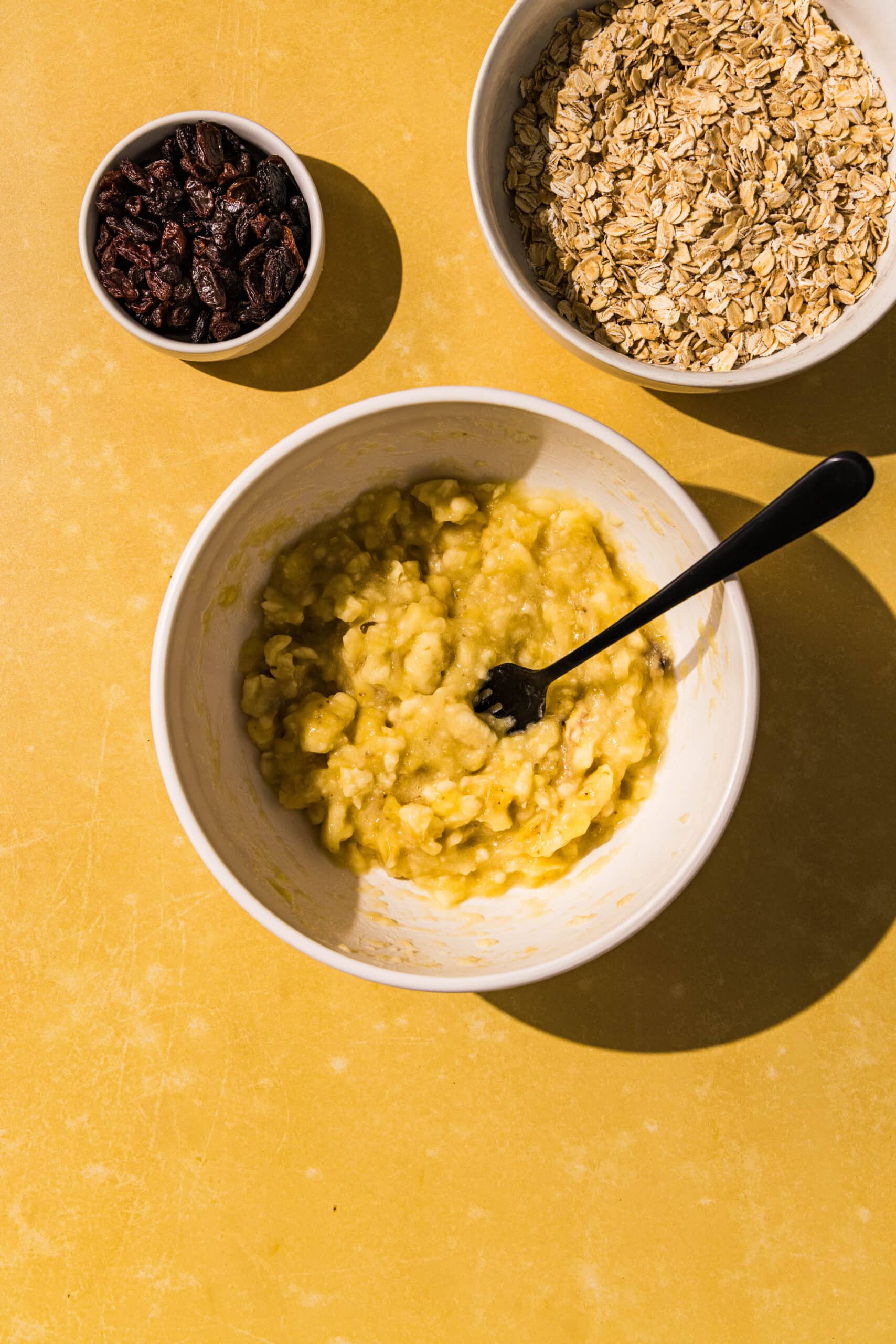 A white bowl of mashed banana with a black fork in it. Above it is two smaller white bowls of rolled oats and raisins.