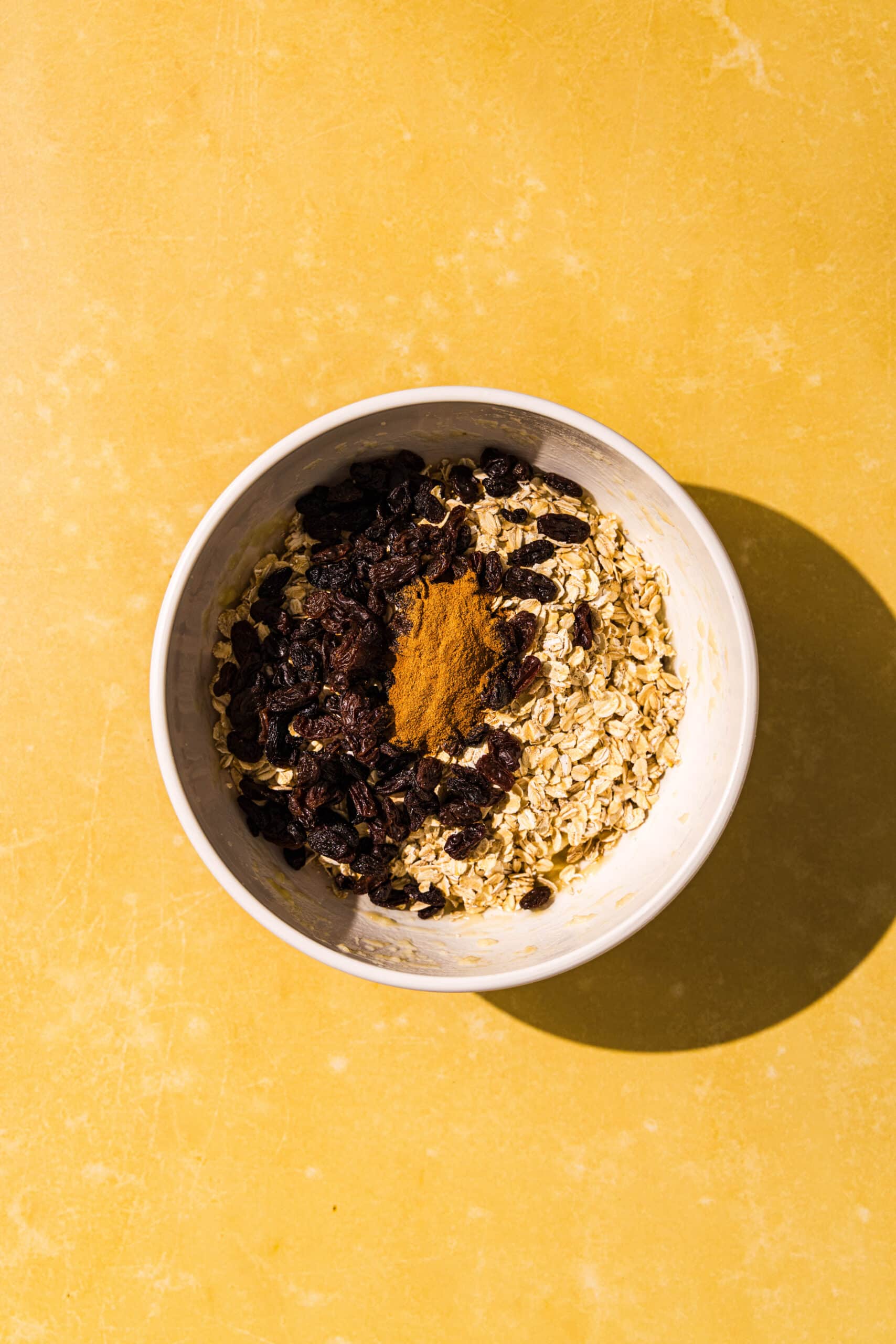 A white bowl of mashed banana with rolled oats, raisins, and cinnamon thrown in on top and ready to be mixed.