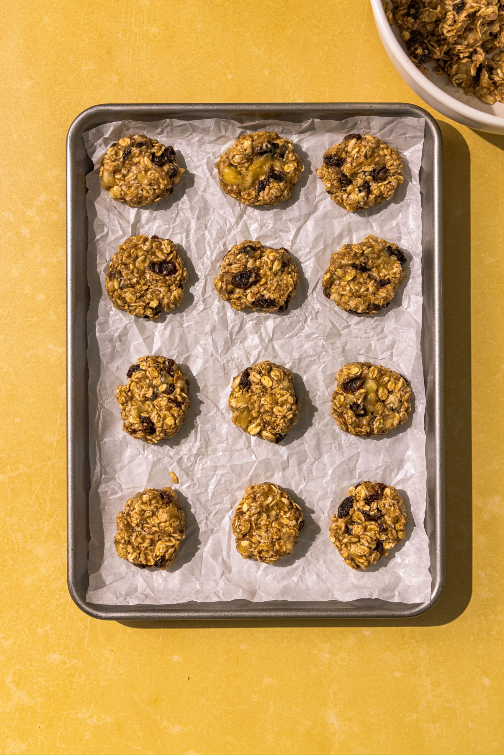 A silver pan lined with parchment paper and filled with 12 banana oatmeal bites lined up ready to be baked. In the left corner is part of a white bowl filled with the banana oat bite batter.