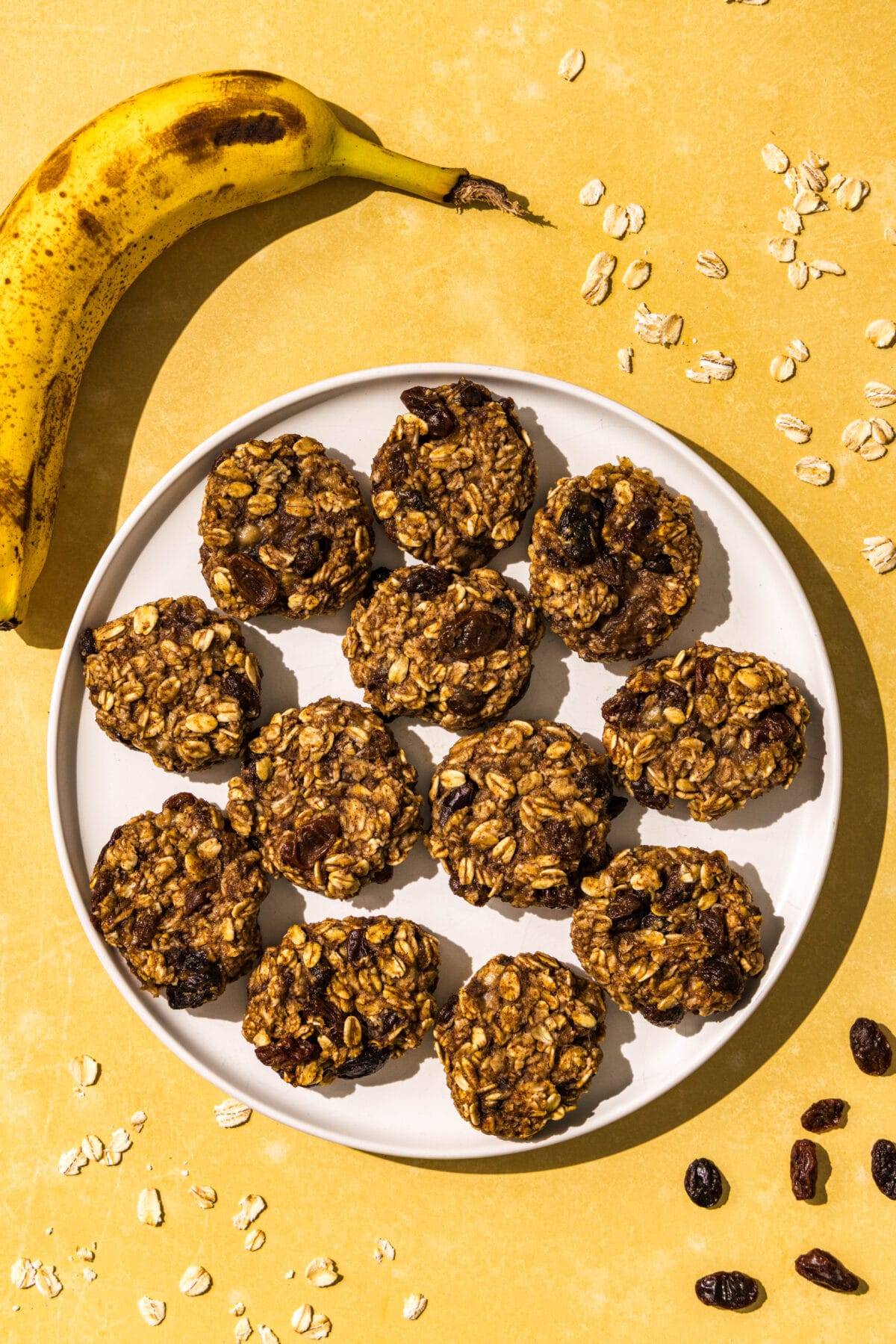 A white plate of 3-ingredient banana oatmeal bites on it. Around the plate sprinkled on the counter is raisins and oats. In the top left next to the plate is a ripe banana on the counter.