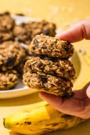 Three 3-ingredient oatmeal banana bites stacked together and being held by a white hand from the right of the image. Below the bites is a banana and in the background is a white plate with oatmeal bites on it and some oats sprinkled on the counter.