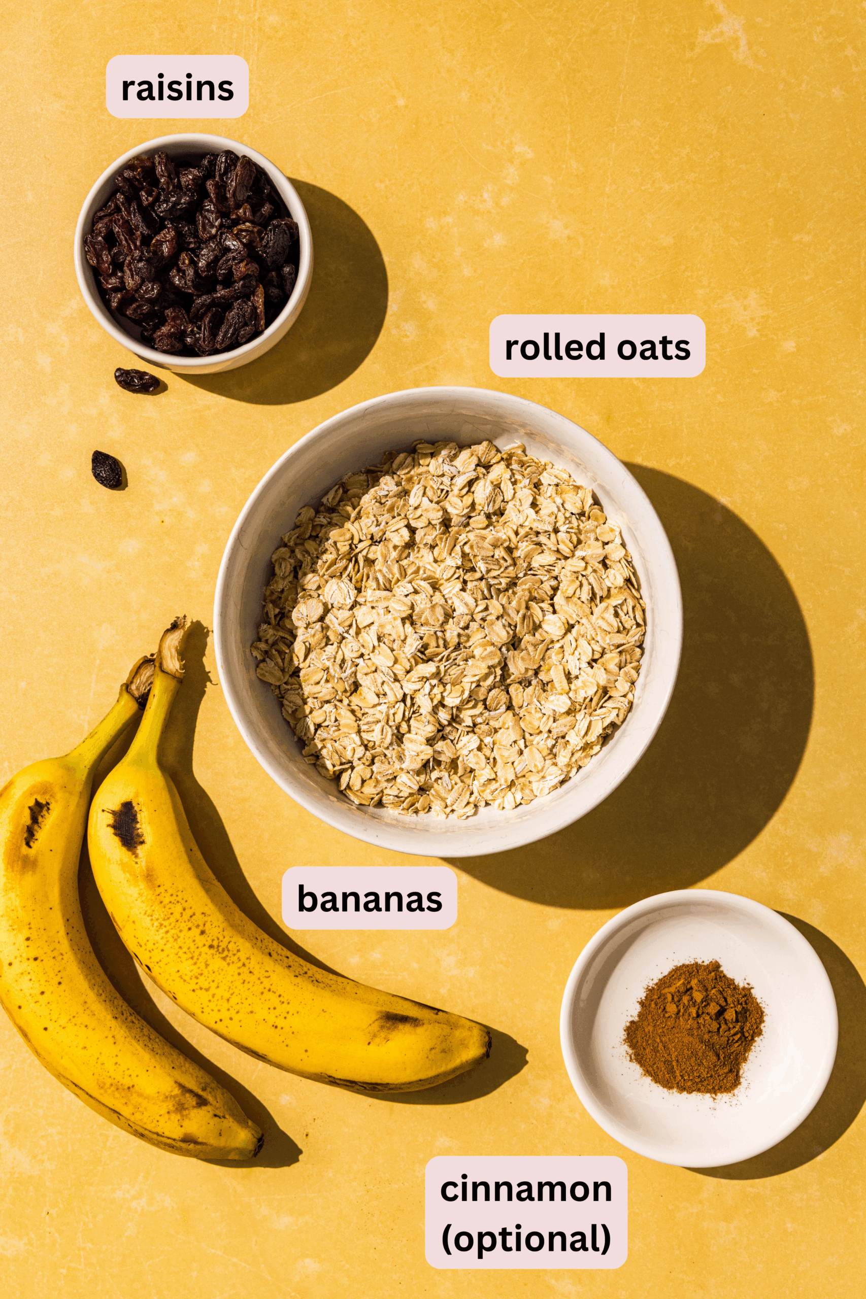 Ingredients needed to make 3-ingredient banana oatmeal bites laid out on a yellow counter and labeled including: raisins, rolled oats, banana, and cinnamon.
