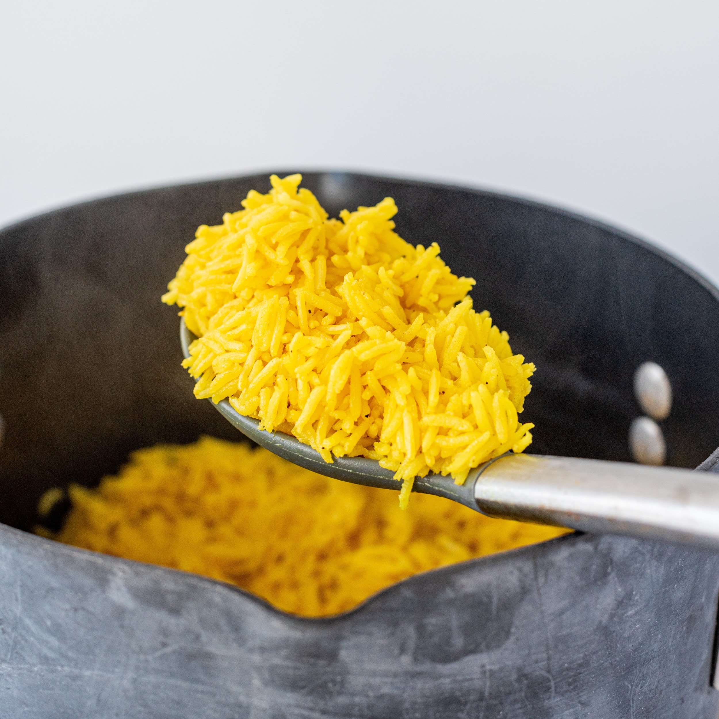 A black pot filled with yellow rice with a silver spoon above it holding a scoop of rice.
