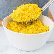 A white bowl of yellow rice with a spoon above it holding a large scoop of rice. Behind the bowl is a silver pot on a cork board.