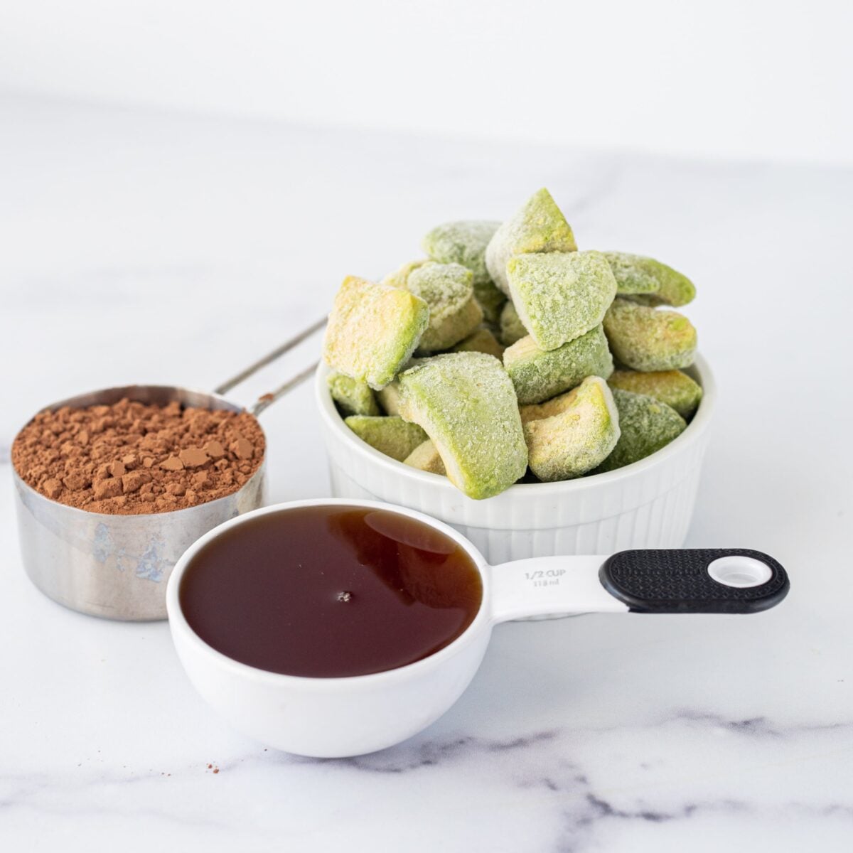 Two measuring cups with cacao powder and maple syrup and a bowl of frozen avocado cubes.