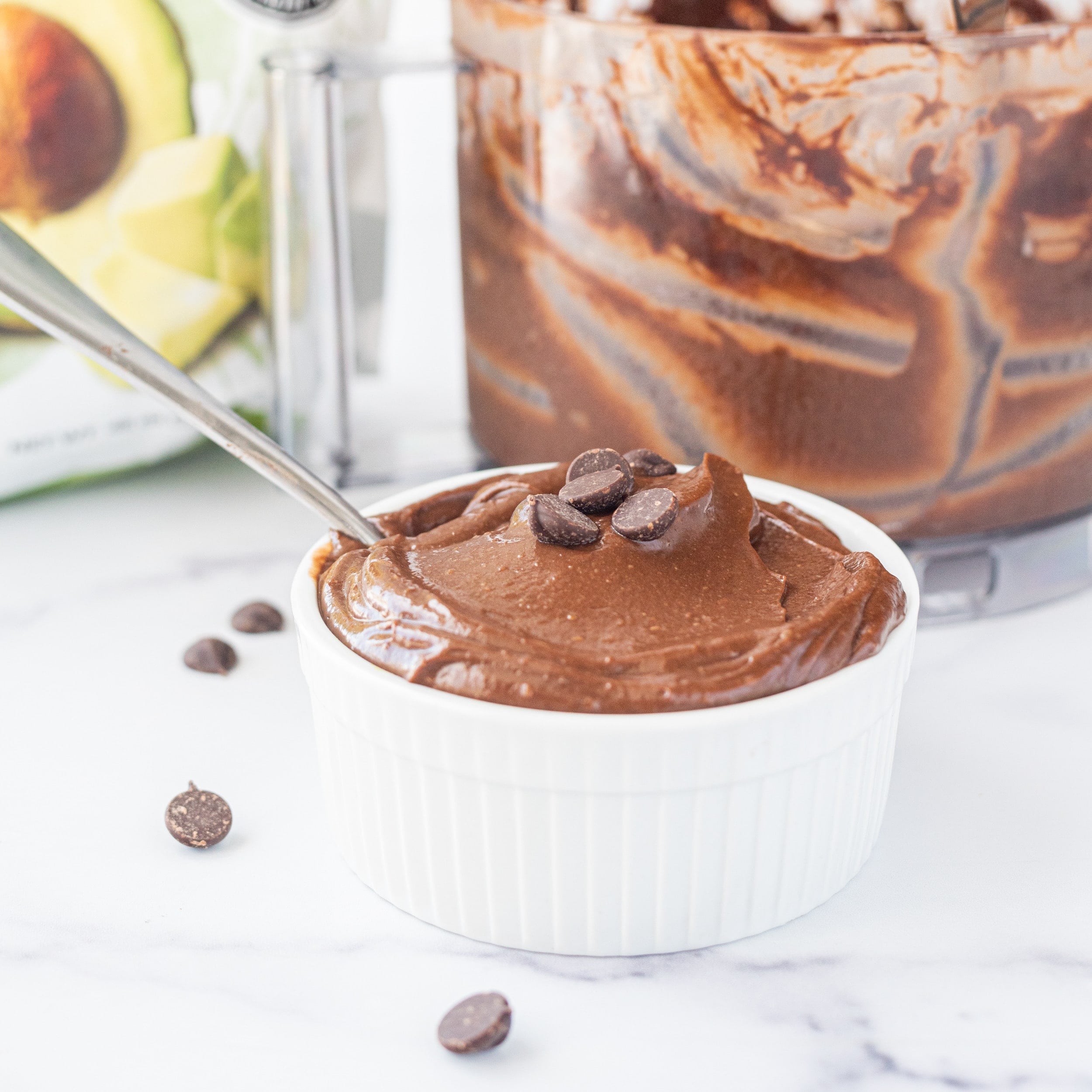 Half of an avocado, a bowl of chocolate pudding with a spoon in it, and a food processor with chocolate pudding in it.