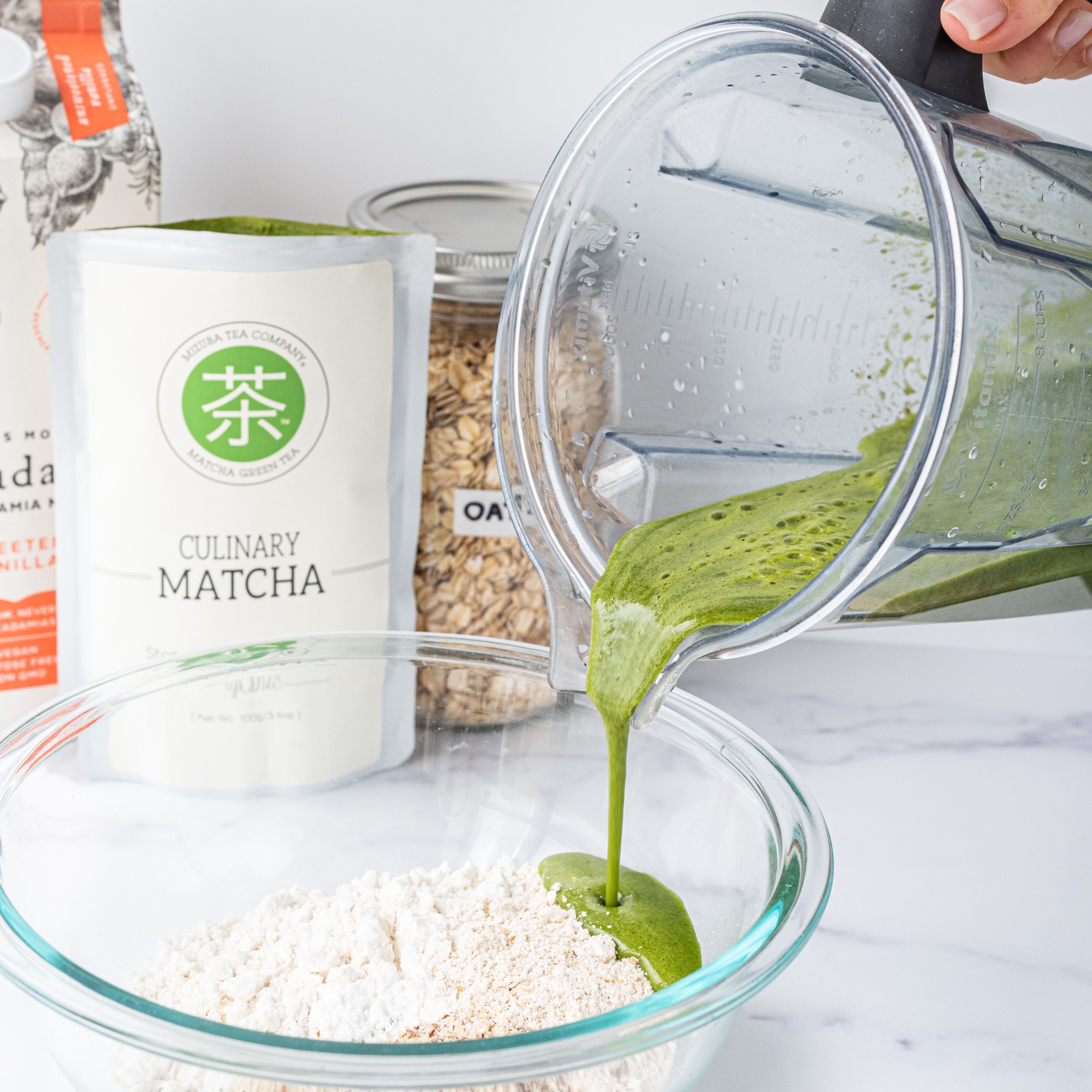 A glass bowl filled with flour and a blender pouring in a green liquid mixture into the glass bowl.