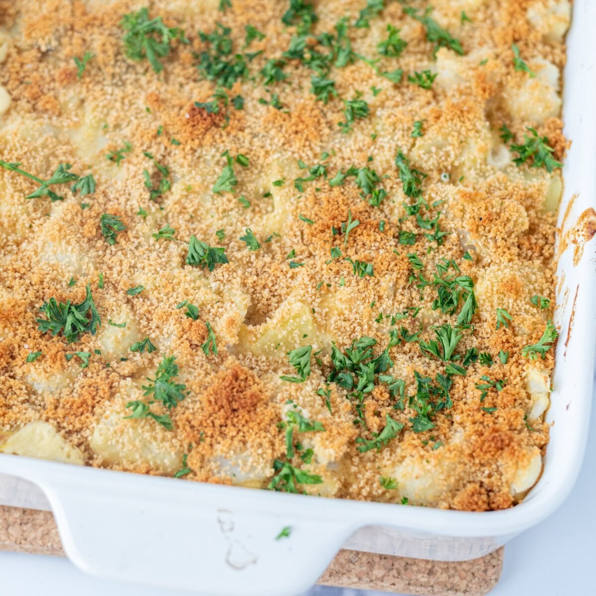Close up of a Cheesy Vegan Cauliflower Potato Casserole in a white casserole dish on a cork pot holder. The casserole is topped with toasted breadcrumbs and fresh parsley.
