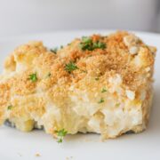A white plate with a silver spatula on it holding a square of Cheesy Vegan Cauliflower Potato Casserole garnished with breadcrumbs and fresh parsley.