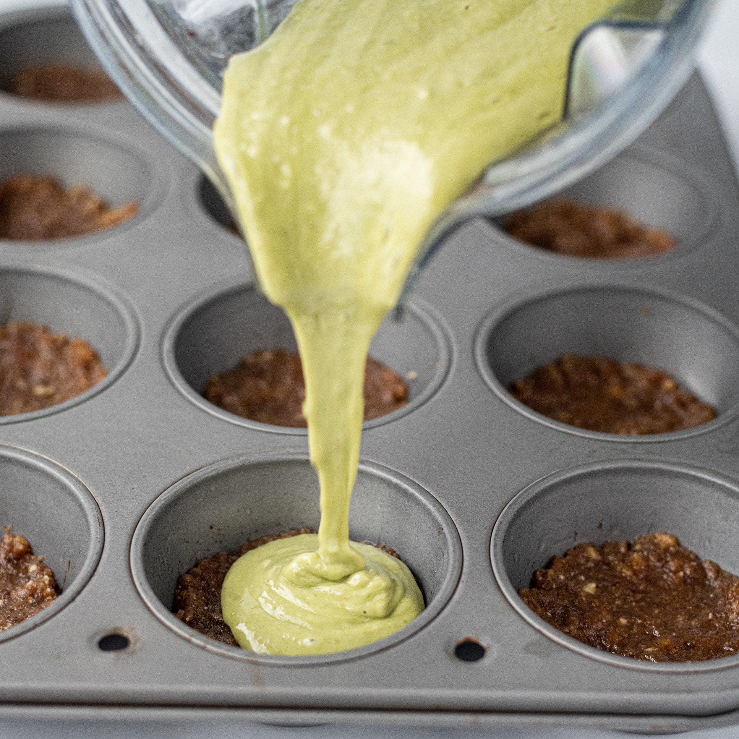 A muffin tin with crust in the muffin cups and a blender pouring matcha cheesecake batter into one of the cups.