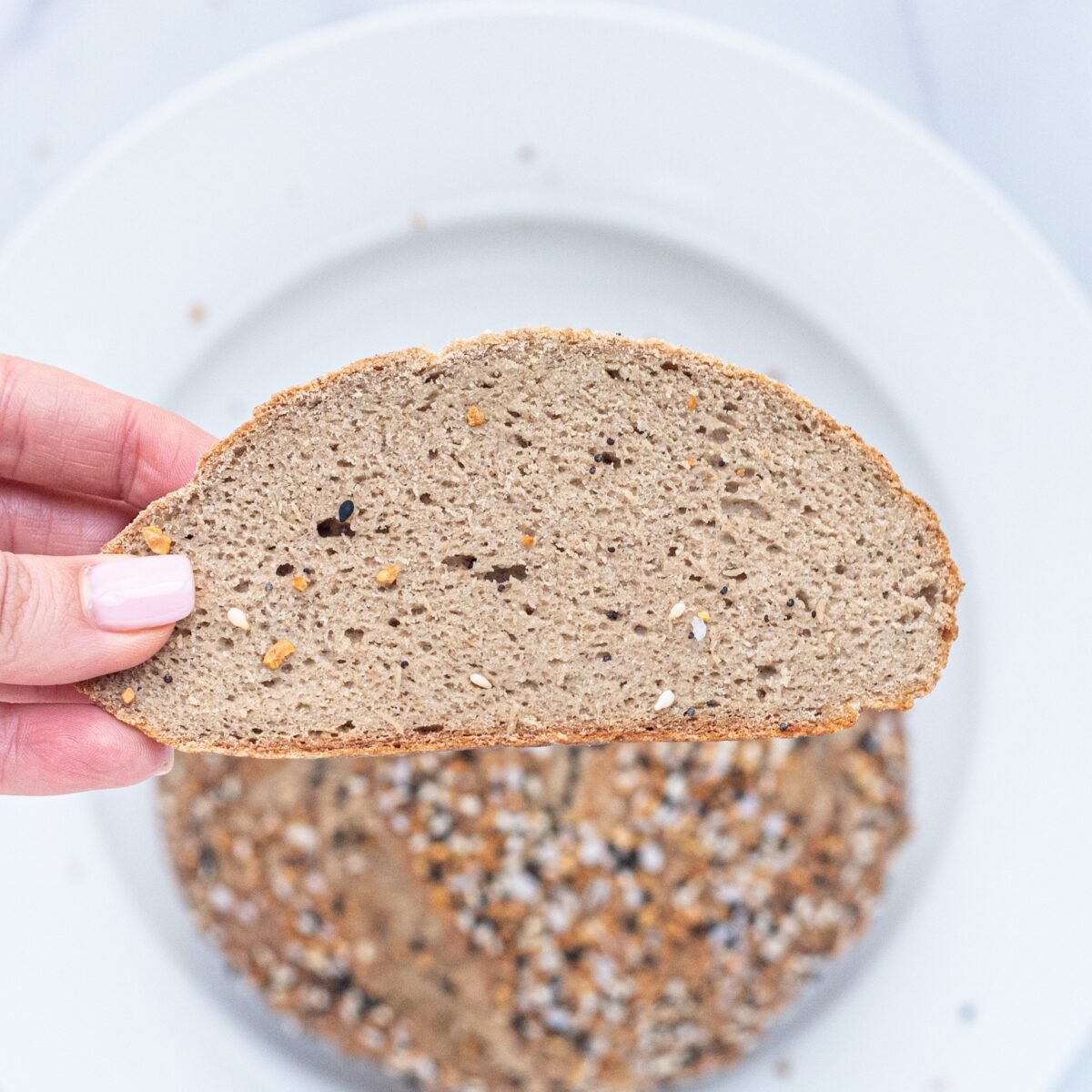 A white plate with a round loaf of bread on it in the background and a slice of bread being held by a white hand with pink fingernails in the foreground.