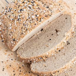 A loaf of bread on a wood cutting board with three slices cut off on the end with everything seasoning on top.