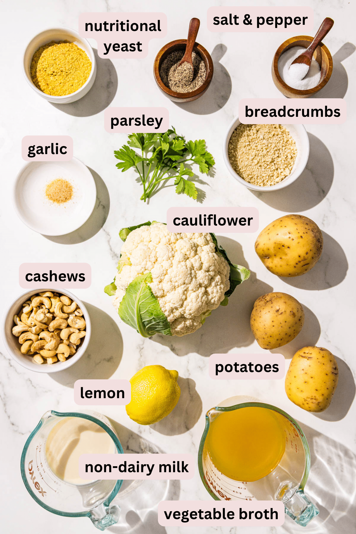 Ingredients laid out on a marble countertop in bowls and labeled including salt and pepper, nutritional yeast, parsley, breadcrumbs, garlic powder, a whole head of cauliflower, potatoes, cashews, a lemon, vegetable broth, and non-dairy milk.