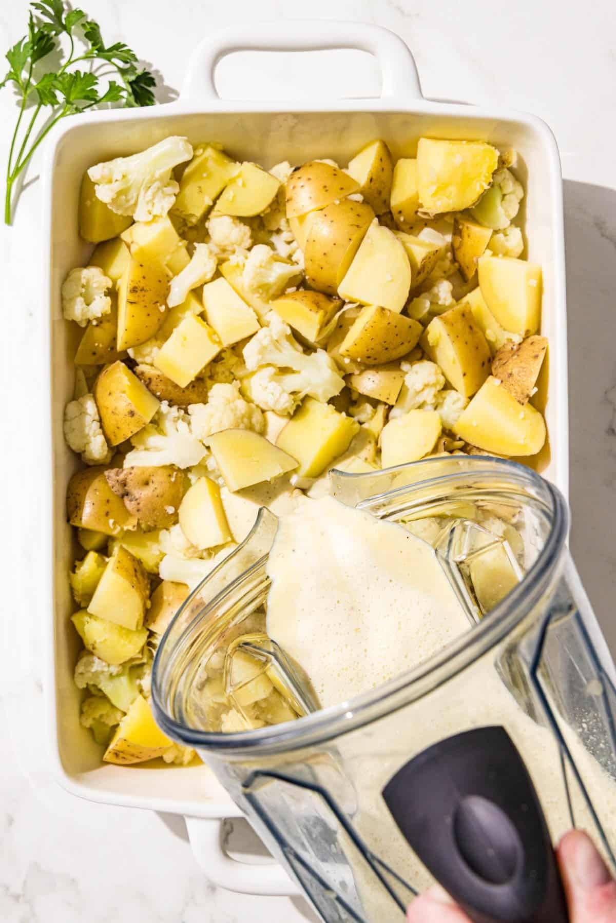A white 9x13 casserole dish filled with steamed potatoes and cauliflower and a clear plastic blender pouring a white sauce over the casserole.