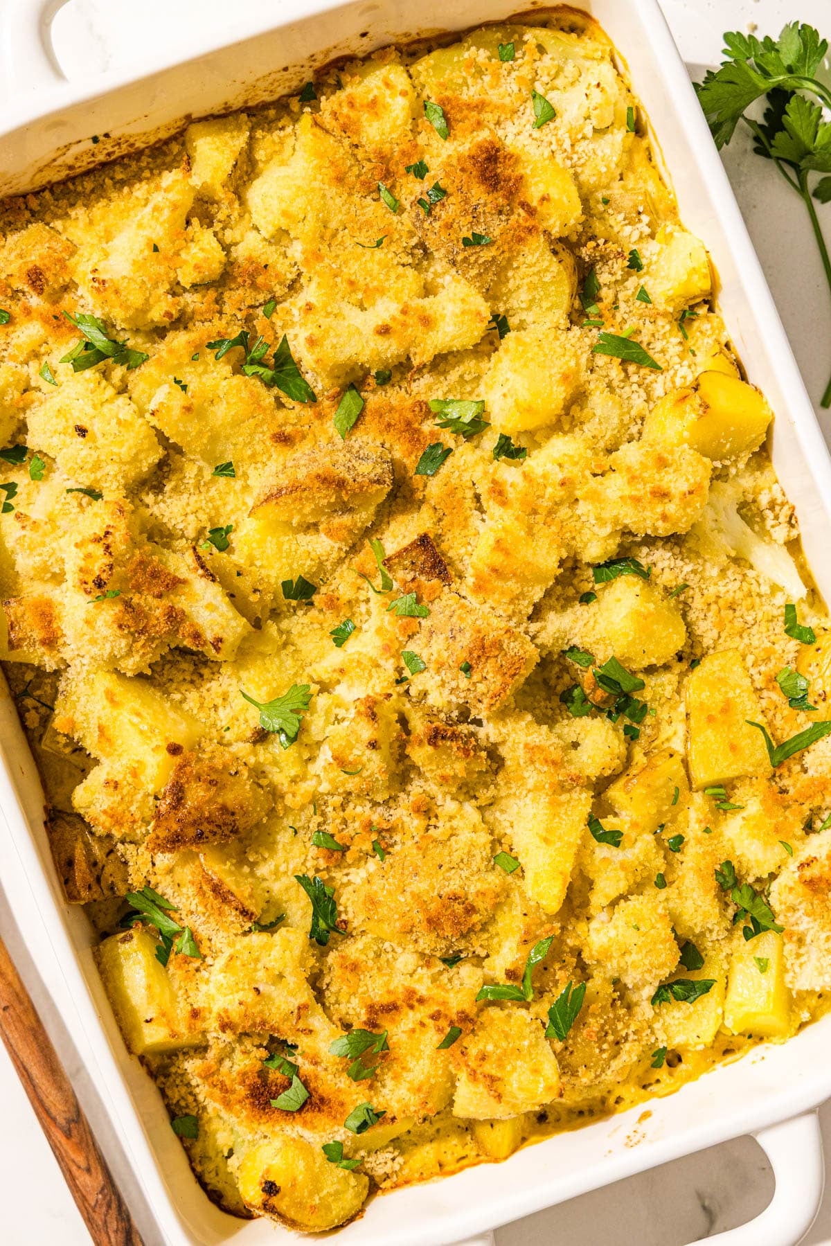 A white glass casserole dish filled with cauliflower potato casserole topped with breacrumbs and parsley. Beside the dish is a wooden spoon and a sprig of parsley.