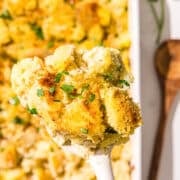 A spatula holding a piece of the cauliflower potato casserole over the white casserole dish with remaining casserole and a wooden spoon.