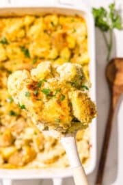 A spatula holding a piece of the cauliflower potato casserole over the white casserole dish with remaining casserole and a wooden spoon.