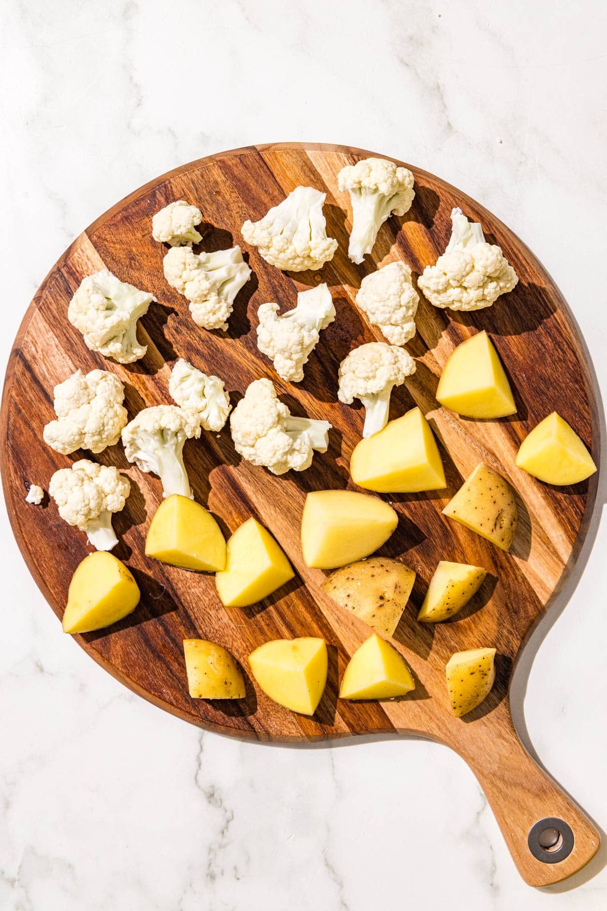 A round wooden cutting board with chopped cauliflower and potato on it.