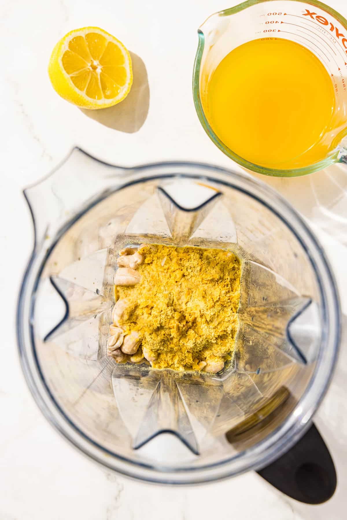 A blender filled with cashews and nutritional yeast. Beside the blender is half of a lemon and a cup of vegetable broth.