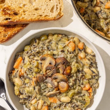 A bowl of creamy wild rice soup topped with mushrooms next to two pieces of toasted bread, a pot with remaining soup, and two metal spoons.