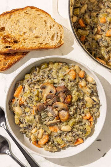 A bowl of creamy wild rice soup topped with mushrooms next to two pieces of toasted bread, a pot with remaining soup, and two metal spoons.